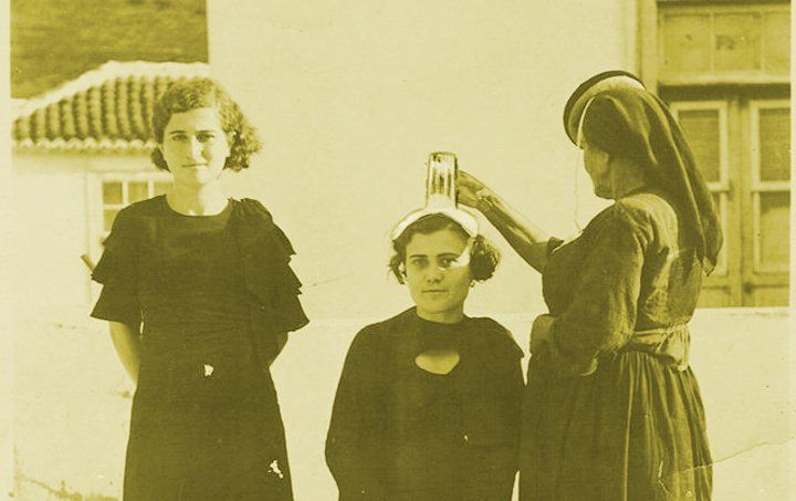 Santiguadoras de canarias. Tres mujeres, dos de pie y una sentada en medio. Una de las mujeres de pie le coloca un vaso en la cabeza a la que está sentada